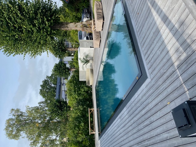 Modern pool med grått trädäck, overflow-kant och välskött trädgård – installation i Mälardalen